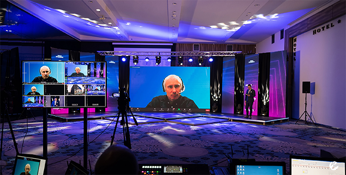 A live event broadcast with a man on a large screen presenting on a topic and several monitors around playing the same broadcast.