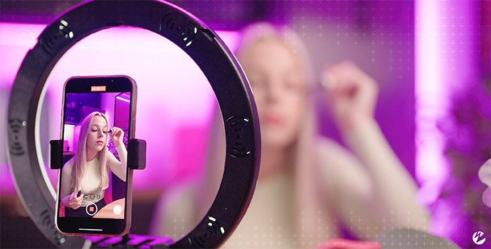 A woman applying makeup for a video tutorial using a ring light and tripod for her smartphone to capture a recording.
